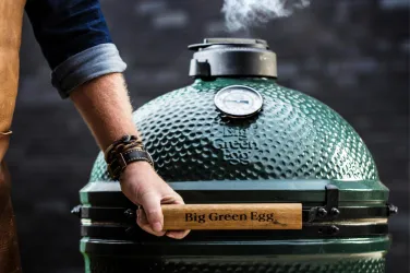 Ein Big Green Egg Grill wird von einer Person mit einer Hand bedient. Rauch steigt aus dem Grill auf, was auf das Grillen von Lebensmitteln hinweist.