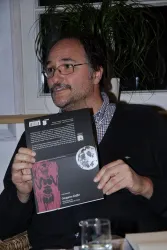 Ein Mann hält ein Buch in der Hand und zeigt die Rückseite. Im Hintergrund sind Pflanzen und eine Fensterbank zu sehen.