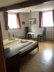 Ein einfaches Schlafzimmer mit einem großen Bett und weißen Bettdecken. Zwei Stühle und ein Tisch ergänzen die Einrichtung, während der Raum mit roten Vorhängen dekoriert ist.