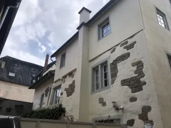 Ein altes, charmantes Haus mit einem hellen Putz und einigen unregelmäßigen Steinelementen. Der Himmel ist teilweise bewölkt, was dem Bild eine stimmungsvolle Atmosphäre verleiht.