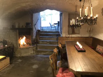 Ein gemütlicher Keller mit einem großen Holztisch und Stühlen. Im Hintergrund brennt ein Kamin und es gibt eine Treppe, die nach draußen führt.