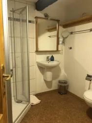 Ein einfaches Badezimmer mit einer Dusche, einem Waschbecken und einemWC. Weiße Fliesen und ein Holzregal sorgen für eine helle Atmosphäre.