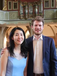 Ein Paar steht in einer Kirche, im Hintergrund sind Orgelpfeifen zu sehen. Die Frau trägt ein hellblaues Kleid, während der Mann einen dunklen Anzug trägt.