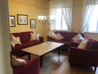 Ein gemütlicher Wohnraum mit roten Sofas und einem Holztisch in der Mitte. Große Fenster lassen viel Licht herein und schaffen eine einladende Atmosphäre.