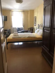 Ein einfaches Schlafzimmer mit einem großen Bett und zwei Stühlen. Das Licht der Fenster schafft eine angenehme Atmosphäre.