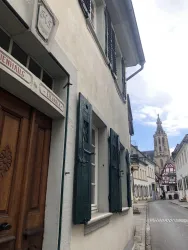 Eine alte Straße mit charmanten Häusern und grünen Fensterläden. Im Hintergrund ist ein Kirchturm zu sehen.