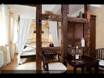 Ein stilvolles Schlafzimmer mit einem Bett, umgeben von eleganten Vorhängen. Ein kleiner Tisch mit zwei Gläsern Roséwein ergänzt die gemütliche Atmosphäre.