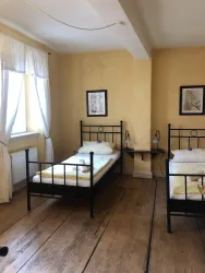 Ein einfaches Zimmer mit zwei Einzelbetten und einem großen Fenster. Die Wände sind in einem warmen Gelbton gestrichen und es gibt zwei Bilder an der Wand.