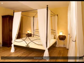 Ein romantisches Schlafzimmer mit einem Himmelbett und sanften, weißen Vorhängen. Die Wände sind in warmen Gelbtönen gehalten, und es gibt rustikale Holzmöbel.