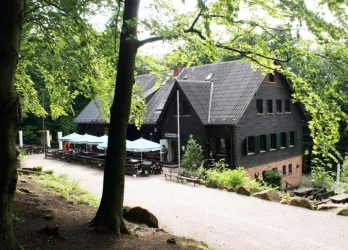 Ein stilvolles Holzgebäude umgeben von Bäumen im Wald. Vor dem Gebäude stehen Tisch- und Sitzmöglichkeiten im Freien.