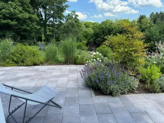 Ein moderner Garten mit bunten Blumen und grünen Sträuchern. Im Vordergrund steht eine Liege auf einem Plattenboden.