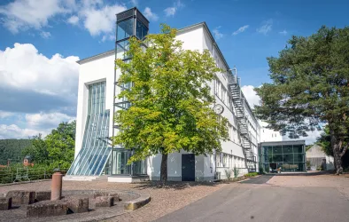 Ein modernes Gebäude mit großen Fenstern und einer Glasfassade ist umgeben von Bäumen. Der Himmel ist klar mit einigen Wolken.
