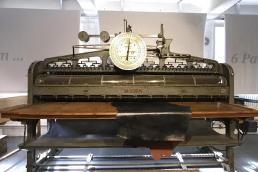 Eine historische Druckmaschine mit einer großen Waage oben. Der Tisch aus Holz ist für die Druckproduktion vorbereitet.