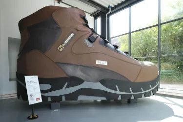 Ein riesiger Wanderschuh aus Kunststoff steht in einem hellen Raum. Die große Skulptur ist mit einer detaillierten Textur und Farbgebung gestaltet.