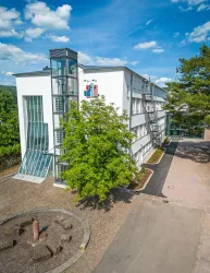 Ein modernes Gebäude mit großen Fenstern und einer gläsernen Treppe. Umgeben von Bäumen und einer klaren Aussicht.