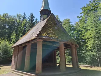 Eine kleine, rustikale Kapelle mit einem grünen Dach und Säulen. Sie steht inmitten eines dichten Waldes.