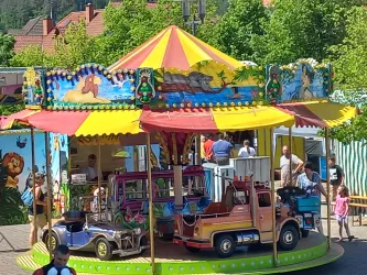 Ein bunter Karussell mit kleinen Autos, umgeben von grünen Bäumen. Viele Kinder und Erwachsene stehen darauf und genießen die fröhliche Atmosphäre.