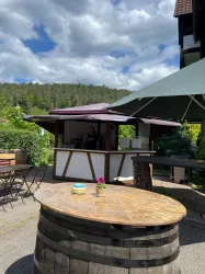 Ein schöner Kiosk mit einer Holzterrasse und einem großen Sonnenschirm. Im Hintergrund sind Bäume und ein blauer Himmel zu sehen.