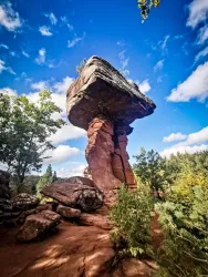 Ein beeindruckender Felsen mit einem großen, flachen Stein oben. Umgeben von Bäumen und einem klaren blauen Himmel.