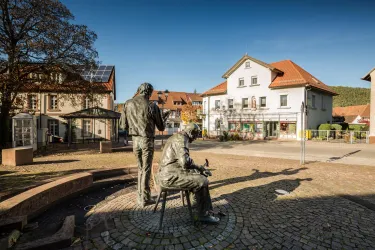 Eine Statue von zwei Männern steht auf einem Platz. Im Hintergrund sind Häuser mit roten Dächern und ein klarer Himmel zu sehen.