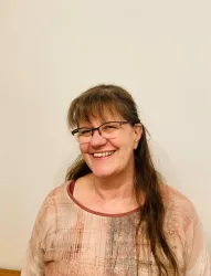 Eine lächelnde Frau mit langen Haaren steht vor einer neutralen Wand. Sie trägt eine gepunktete Bluse und Brille.