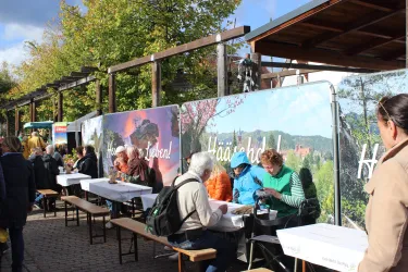 Ein belebter Außenbereich mit Tischen und Bänken, an denen Menschen sitzen und essen. Im Hintergrund sind großformatige Bilder und eine schöne Herbstlandschaft zu sehen.