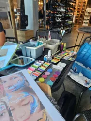 Ein Tisch mit verschiedenen Gesichtsmalutensilien, darunter Farben, Pinsel und eine Anleitung. Im Hintergrund sind Schuhe in einem Geschäft zu sehen.