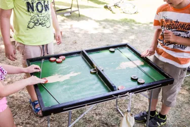 Kinder spielen an einem Tisch mit einem Spiel, das eine grüne Spielfläche hat. Es gibt mehrere Spielsteine, die sie bewegen.