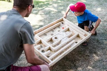 Ein Erwachsener und ein Junge spielen mit einem Holzspielzeug auf dem Rasen. Das Spielbrett zeigt ein Labyrinth, das sie gemeinsam erkunden.
