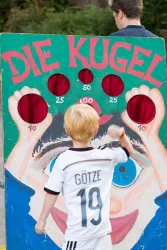 Ein Junge wirft einen Ball auf eine Zielscheibe mit verschiedenen Punkten. Im Hintergrund steht ein Erwachsener.