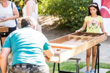 Ein Kind spielt an einem Holzspieltisch im Freien. Ein Erwachsener steht davor und zielt auf ein Spielobjekt.