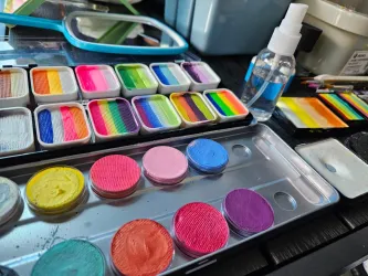 Eine Vielzahl von farbigen Gesichtern und Malutensilien sind auf einem Tisch angeordnet. Es gibt Sprühfläschchen und verschiedene Farbtöpfe in bunten Tönen.