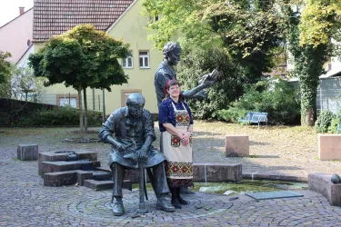Eine Frau steht zwischen zwei Skulpturen in einem Park. Die Skulpturen zeigen zwei Männer, die anscheinend in einer kreativen oder handwerklichen Tätigkeit beschäftigt sind.