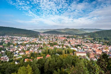 Eine malerische Landschaft mit einem Blick auf eine Stadt umgeben von Hügeln und Wäldern. Der Himmel ist blau mit einigen Wolken.