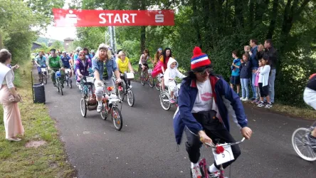 Eine Gruppe von Radfahrern startet bei einem Wettkampf unter einem roten Startbanner. Zuschauer stehen an der Strecke und beobachten das Ereignis.