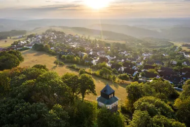 Eine malerische Landschaft mit einem kleinen Dorf im Hintergrund und der Sonne, die über den Horizont aufgeht. Bäume umgeben die Szenerie und schaffen eine ruhige Atmosphäre.
