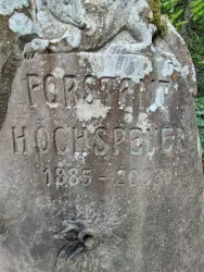 Ein alter Stein mit der Inschrift „Forststadt Hochspeyer 1885 - 2003“. Umgeben von natürlicher Vegetation.
