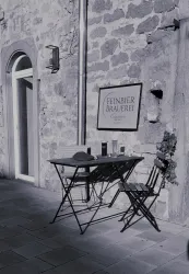 Ein kleiner Tisch mit zwei Stühlen steht vor einer steinernen Wand. An der Wand hängt ein Schild mit der Aufschrift „Einber Schönheit“.