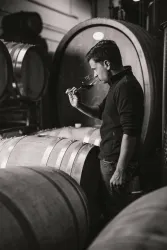 Ein Winzer in einem Weinkeller, der einen Schluck Wein aus einem Glas probiert. Im Hintergrund sind große Holzfässer zu sehen.