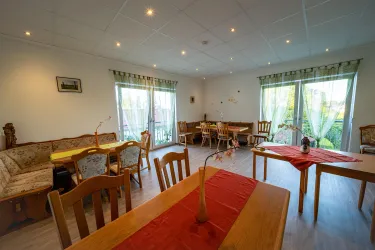 Ein gemütlicher Raum mit Holztischen und Stühlen. Große Fenster sorgen für viel Tageslicht und Blick ins Freie.