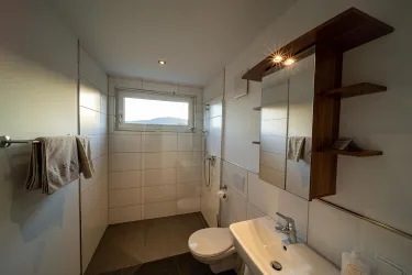 Ein modernes Badezimmer mit weißen Fliesen und einem Fenster. Es gibt eine Dusche, ein Waschbecken und einen Holzregal.