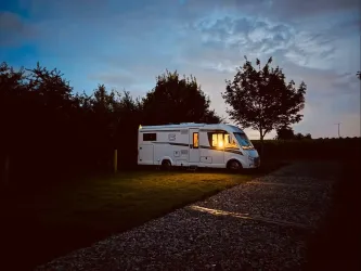 Ein Wohnmobil steht auf einem ruhigen Parkplatz umgeben von Bäumen. Das warme Licht aus dem Fenster strahlt in die Dämmerung.
