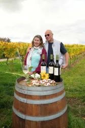Ein Paar steht lächelnd an einem Weinfass in einem Weinberg. Auf dem Fass stehen mehrere Flaschen Wein und eine hübsche Dekoration mit Blumen und Snacks.