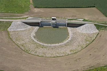 Ein künstlicher Teich mit einem Damm in der Mitte. Umgeben von landwirtschaftlichen Feldern und grünen Rändern.