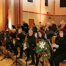 Eine große Musikkapelle spielt in einem festlich dekorierten Raum. Die Musiker tragen schwarze Kleidung und haben hübsche Blumenarrangements an ihren Notenständern.