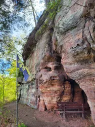 Eine beeindruckende Felswand mit bunten Schichten und einer Sitzgruppe zum Ausruhen und Picknicken. Nebenan steht eine Fahne, die den Ort kennzeichnet.