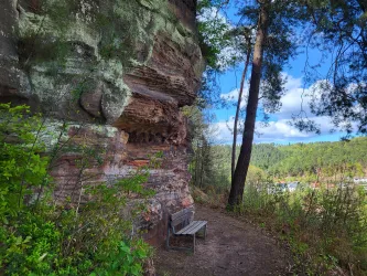 Ein ruhiger Pfad, der an einer Felswand mit einer Holzbank davor vorbeiführt. Im Hintergrund sind Bäume und ein blauer Himmel zu sehen.