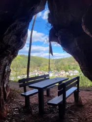 Ein Blick aus einer Höhle auf eine malerische Landschaft mit einem Tisch und Bänken im Vordergrund. Der Himmel ist blau und die Natur erblüht.