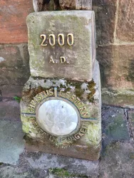 Ein alter Stein mit der Inschrift "2000 A.D." und einer runden Platte mit der Aufschrift "Christus gestern, heute und in Ewigkeit". Der Stein ist teilweise von Moos bedeckt und steht auf einem Steinboden.