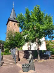 Eine alte Kirche mit einem hohen Turm und einer Statue einer Frau, die einen Korb trägt. Im Hintergrund steht ein großer, grüner Baum.